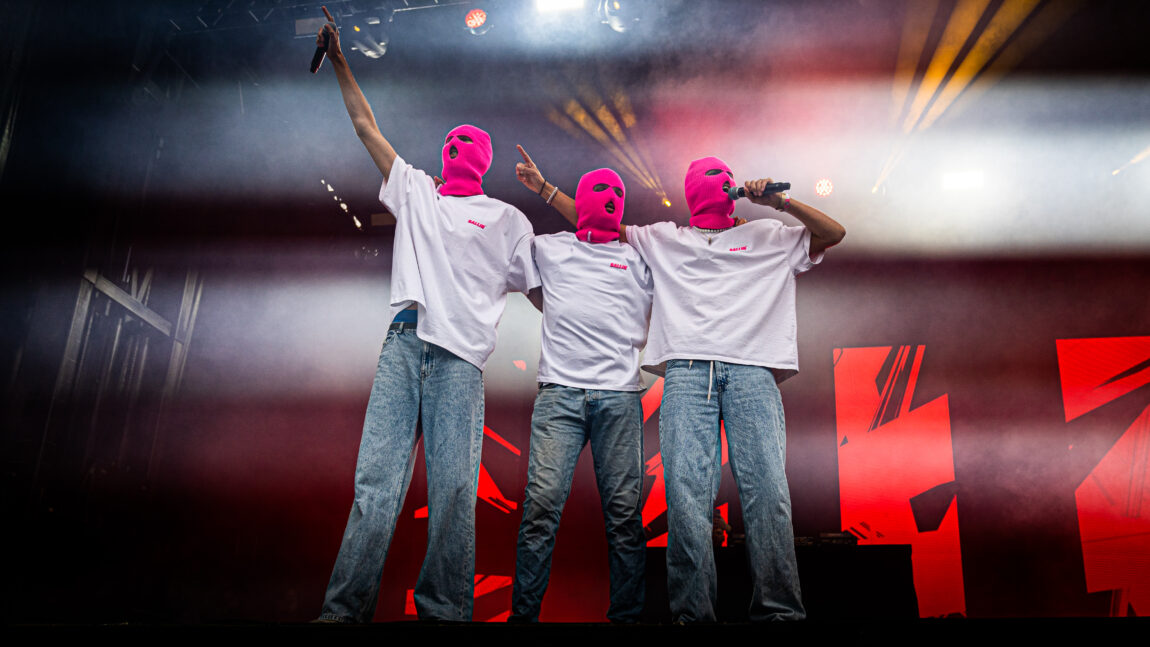 Ballinciaga har på seg hvite t-skjorter, rosa masker og blå jeans. De holder rundt hverandre og synger. 