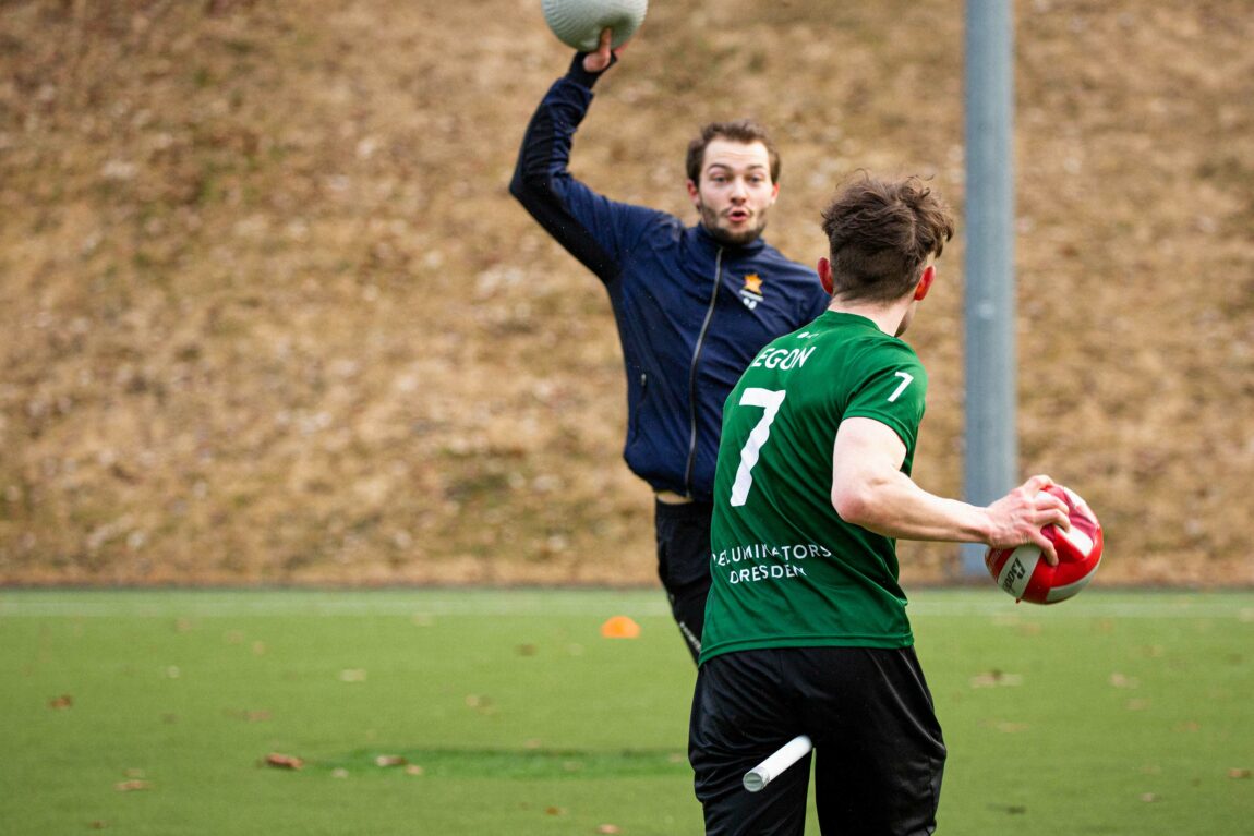 En spiller med fokusert fjes og hevet høyre hånd som holder en ball
og er klar til å skyte. Spilleren vi ser fjeset på er vendt mot en annen spiller som vi kun ser ryggen til. Også sistnevne spiller har en ball i hånden.