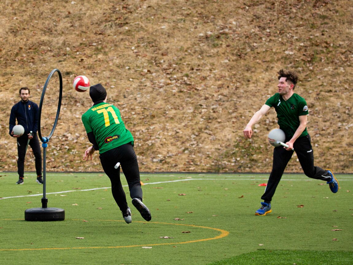 Tre spillere avbildet i fart under treningskamp, utendørs på en fotballbane. Den ene ballen er på vei gjennom en av målringene.