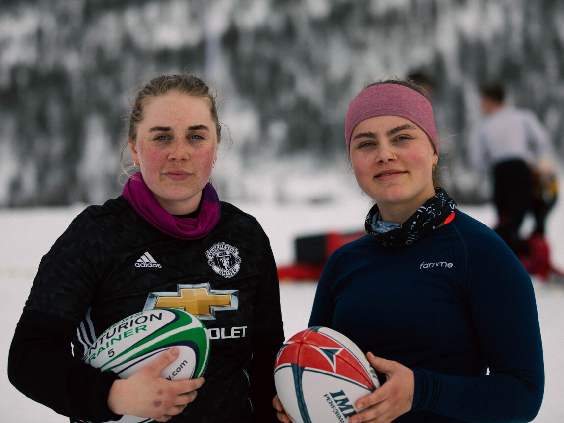 Marte og Guro smilar til kamera med kva sin avlange rugbyball i hendene. Begge har satt opp håret sitt i ein hestehale, har ein buff rundt halsen og har ulltrøyer på. Bak dei er bakken dekka med snø. 