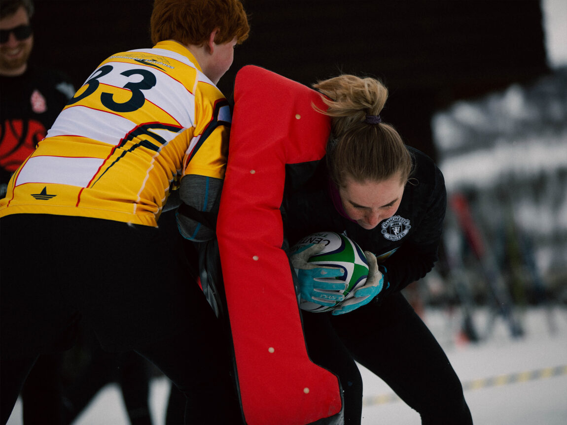 Guro slenger kroppen sin inn i ei raud pute som ein annan spelar held opp. Ho har ballen i hendene og krøker kroppen sin rundt han.