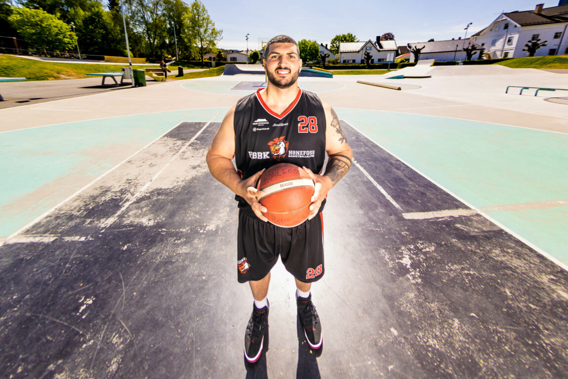 Daniel smiler i kamera mens han holder baketballen mellom begge hender. Bildet er forvridd fordi det er tatt med fisheyelinse, så perspektivet gjør at hendene er veldig store og hele banen bak han synes. 