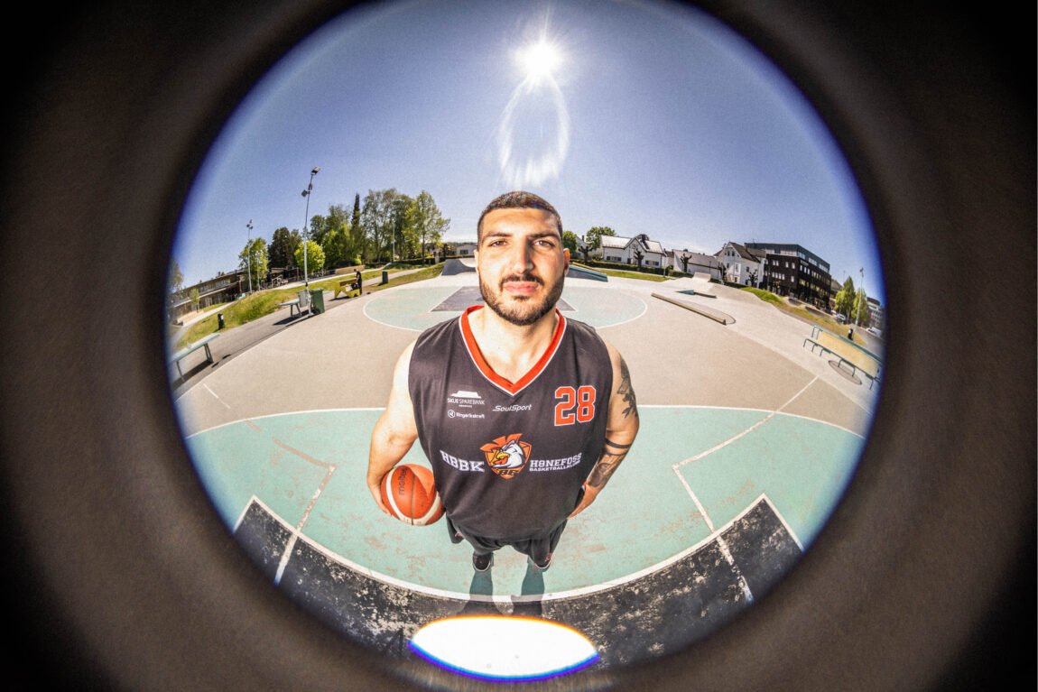 Daniel kikker lurt inn i kamera, som er en fisheyelinse og det ser ut som han kikker inn et kikkhull på en dør. Han holder ballen mellom den ene hånden, og inntil låret på samme side. Man skimter logoen til basketlaget Hønefoss basketballklubb. Sola og himmelen og basketbanen er sirkelformet bak ham - og på siden av linsa er det en svart ramme. 