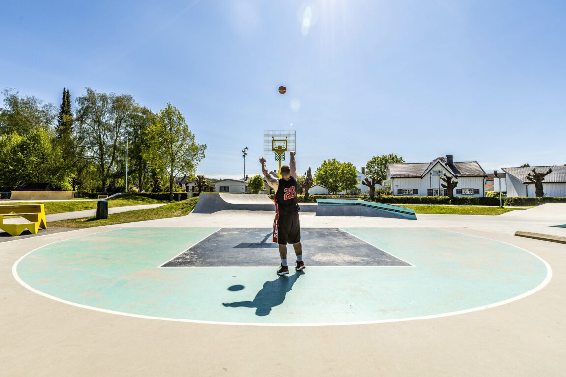 Daniel kaster ballen mot basketballkurven med ryggen til kamera. Man ser tallet 28 i røde. bokstaver printet på ryggen til den sorte singletten. Basketkurven har gult metall og grønn kruv. Rundt han er det solfyllt, man skimter bebyggelse og skatepark. 