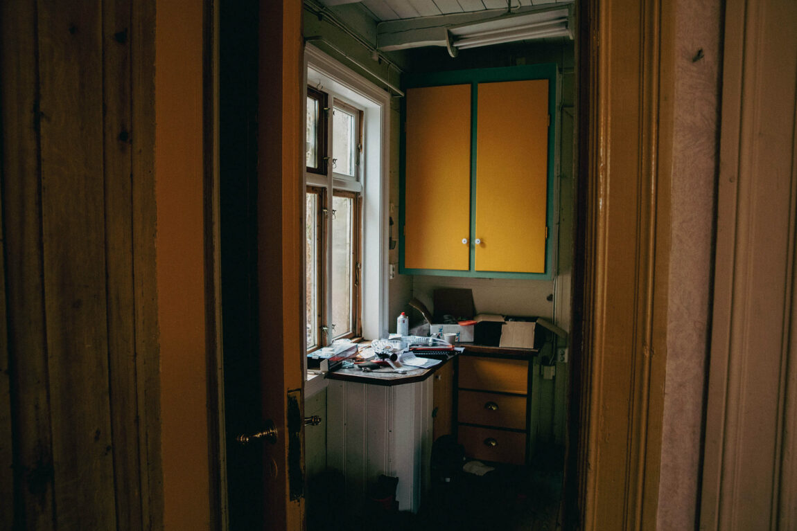Ein kjempeliten kjøkkendisk er fyllt opp av papirer og verktøy. Eit orange og grønt kjøkkenskap heng ved sida av eit vindu. 