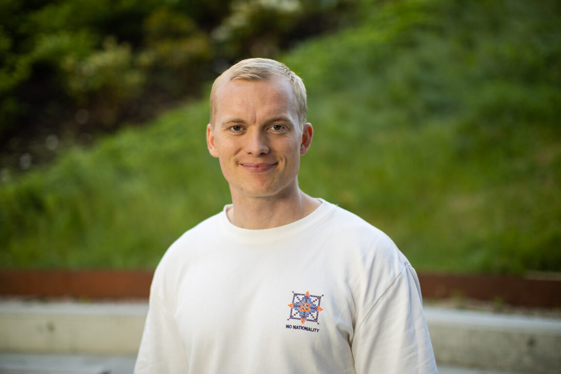 Kasper msiler inn i kamera, med en hvit t-skjorte på. Han har blondt, kort hår. I bakgrunnen er det grønt gress. 