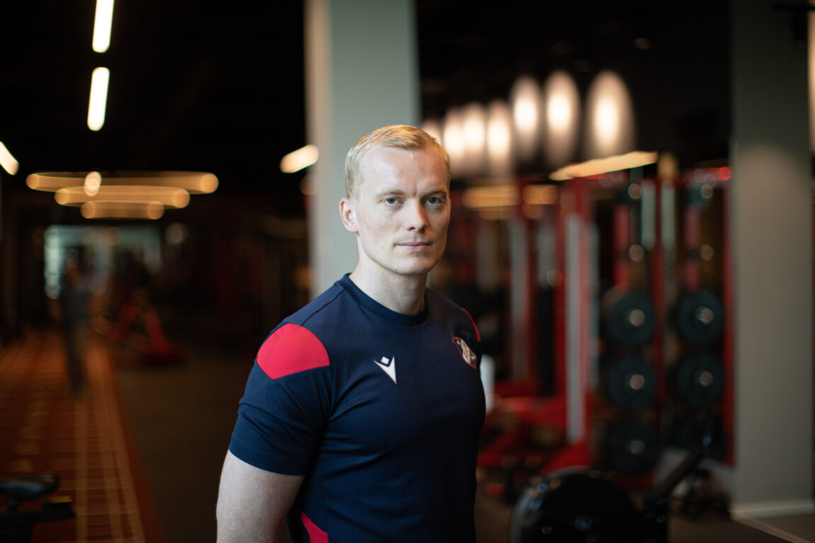 Kasper kikker strengt inn i kamera, med armene bak ryggen. Han har på seg Fram-fotballdrakt, og har blondt kort hår. I bakgrunnen skimter man røde treningsaparater. 