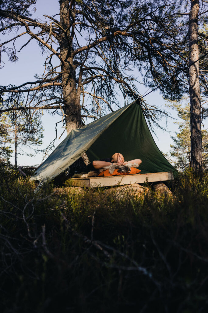 André ligger med hodet på en oransj pute med reinsdyrsskinn oppå. Han viler hodet mot hendene, og albuene hans stikker ut til sidene. Han ligger oppå plattingen, med en militærgrønnfarged tarp hengende over ham - som et trekantet telt. Rundt ham er det trær og grønn lyng. 