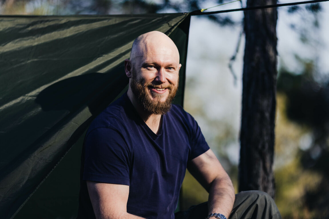 Andre smiler inn i kamera. Han har skjegg og er skalla. Han har på seg en mørkeblå t-skjorte, og sitter foran en mørkegrønn, opphengt tarp. Han lener den ene armen mot låret, hvor han har på mørkegrønn bukse. I bakgrunnen skimter man et bjørketre og blå himmel. 