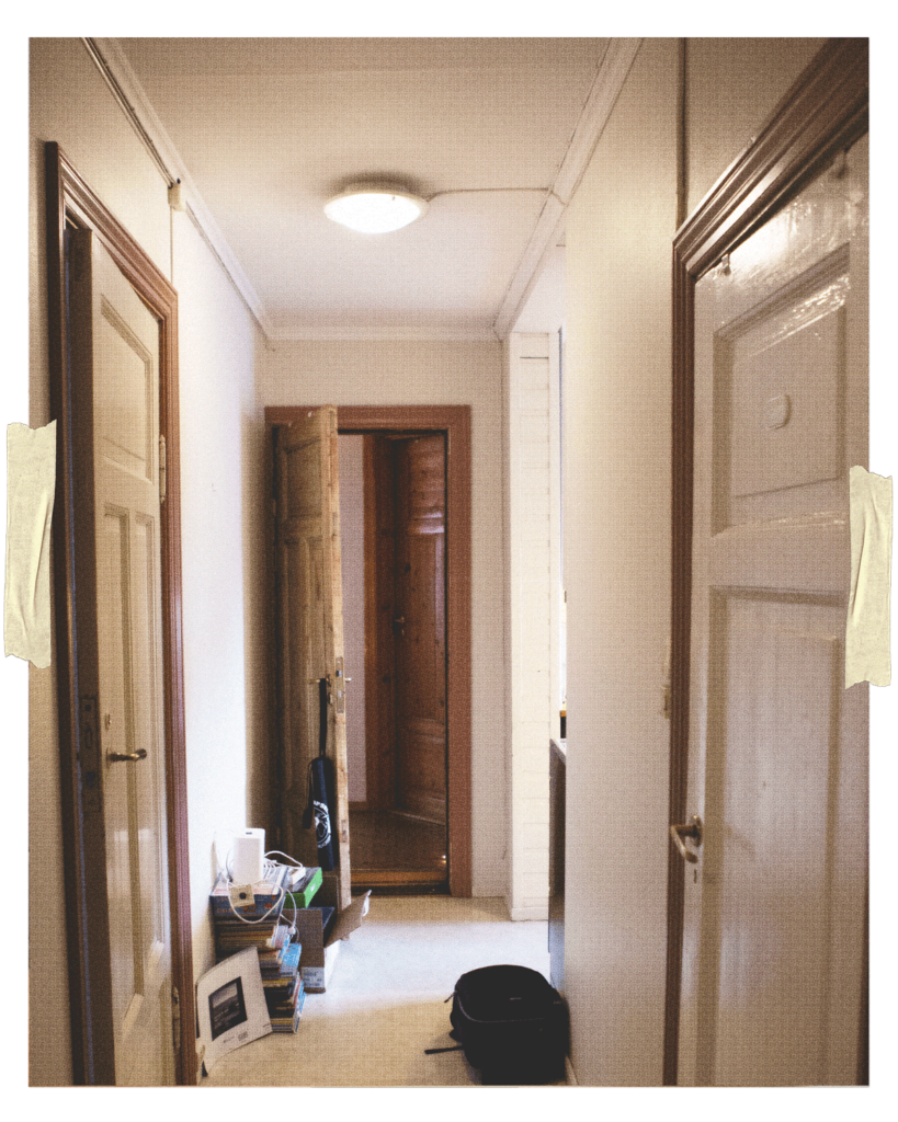 En lang hvitmalt gang med fire synlige tredører er avbildet. Langs den ene veggen står det stablet bøker, en pappeske og noe annet rot. På det ene dørhåndtaket henger et handlenett.