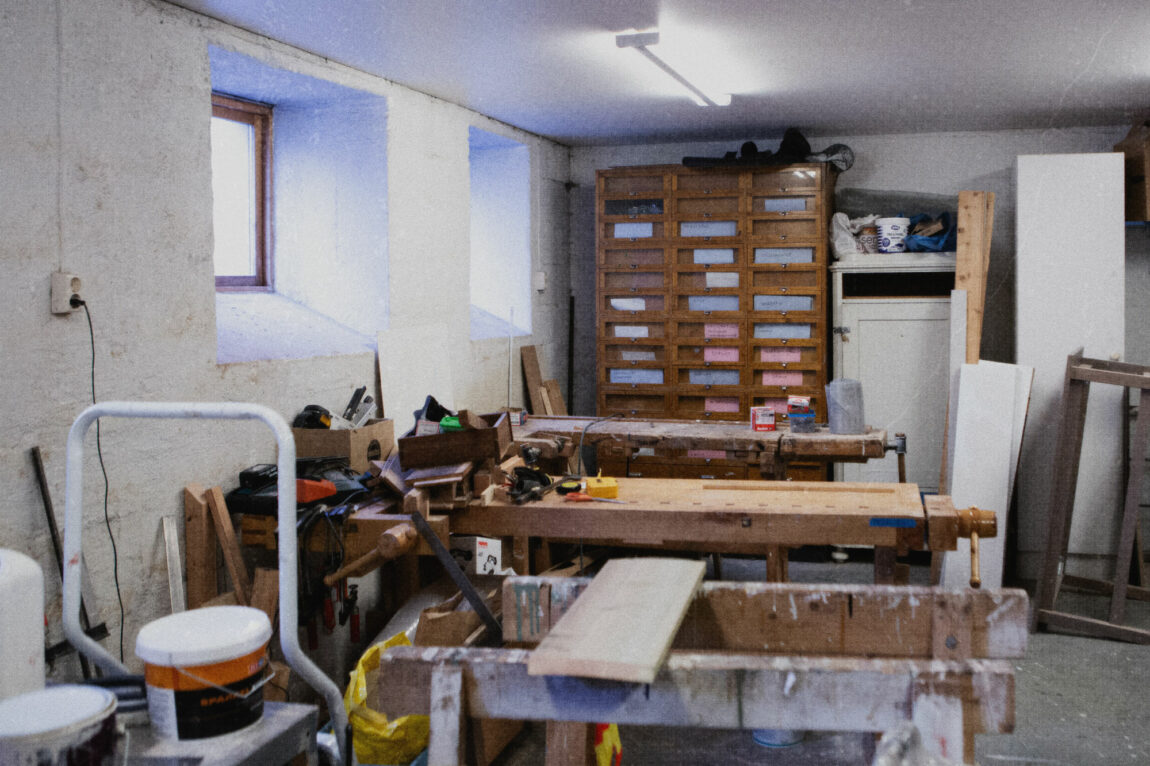 Sløydbenkene er i fokus, ulike hobbyartikler i kasser står oppå benkene. I bakgrunnen viser hyller, og ulike materialer står lent inn mot veggen.