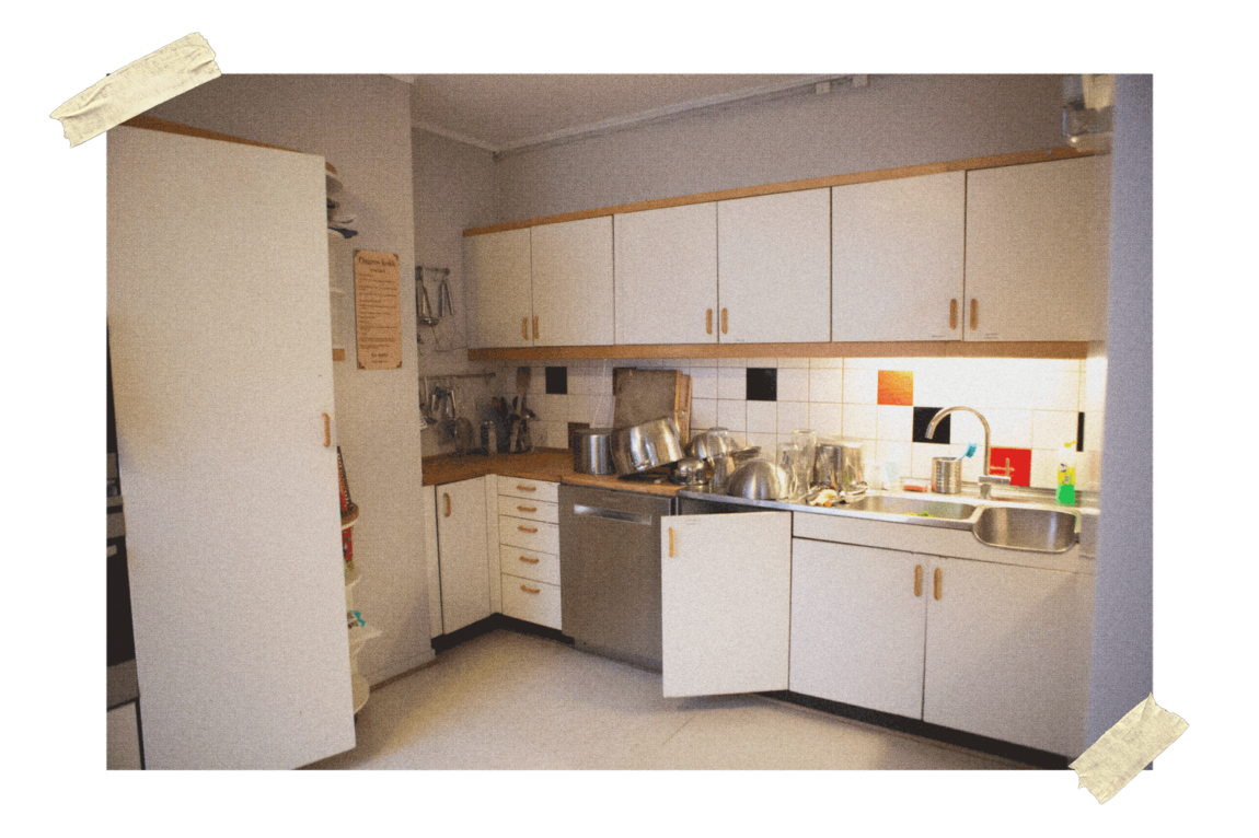 Bildet viser et stort, hvitt og noe retropreget kjøkken med tredetaljer. På benken og ved siden av vaksen står masse kasseroller og annen oppvask til tørk. I bakgrunnen er det hvite fliser, med innslag av orange og svart. 