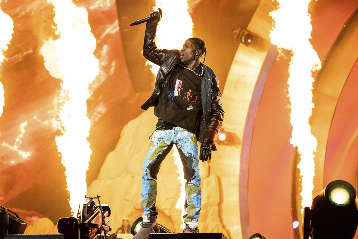 Travis Scott står på scenen med full pyro bak han. Han har på seg en blå bukse, en svart t-skjorte og en svart skinnjakke.