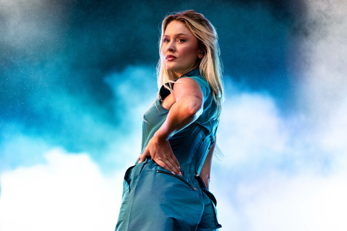 Zara Larsson har på seg en blå bukse og topp. Hun står med rumpa mpt publikum og ser seriøst på publikum