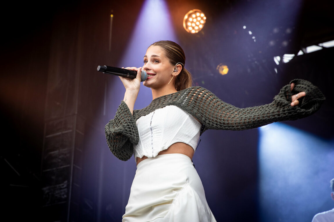 Victoria ikledd en heklet shrug og et hvitt korsett under. Hun står med ene armen ut til siden og i den andre har hun mikrofonen. Hun smiler ut mot publikummet vi ikke ser. 