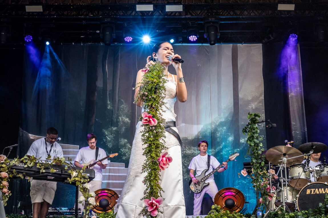 Hillari står på scenen med en mikrofon mot munnen. Mikrofonstativet har masse blomster rundt seg. Bak henne er bandet i full gang. Hun har på seg en hvit kjole.