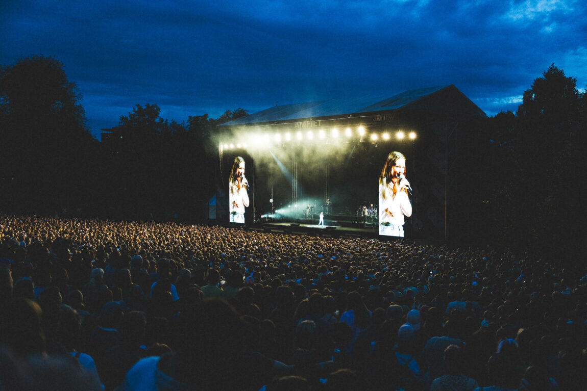 Et hav av publikum står fremfor scenen. På de to skjermene på hver side av scenen ser vi et nærbilde av Sigrid. Det er mørkt, men lyset fra opptredenen lyser over publikum. 