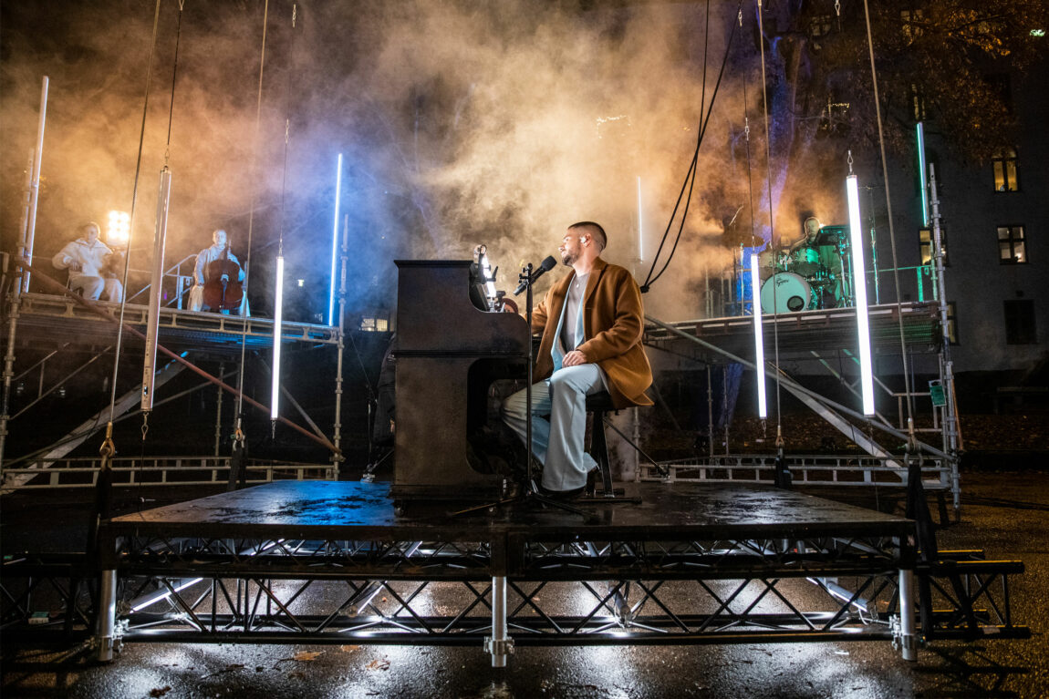 Chris Holsten sitter ved et piano utendørs. Det henger et tau i ryggen hans. Bak ham skimtes musikere på trommer og strykere. Under ham er en plattform som snart skal heises oppover.