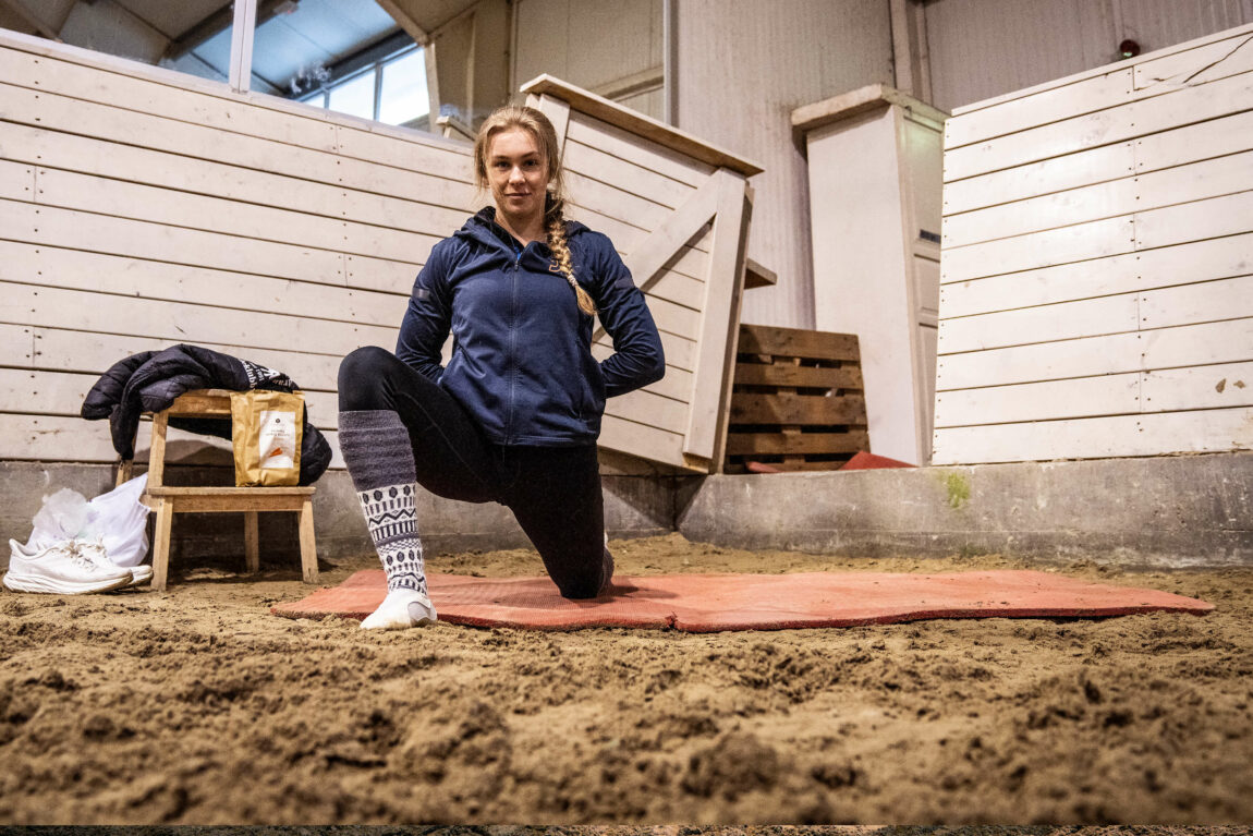 Krstine står med eit kne på ei raud matte og den andre foten framfor seg. Ho har hendene bak ryggen mens ho strekker ut lysken. Me er inne i ridehallen, der underlaget består av sand.