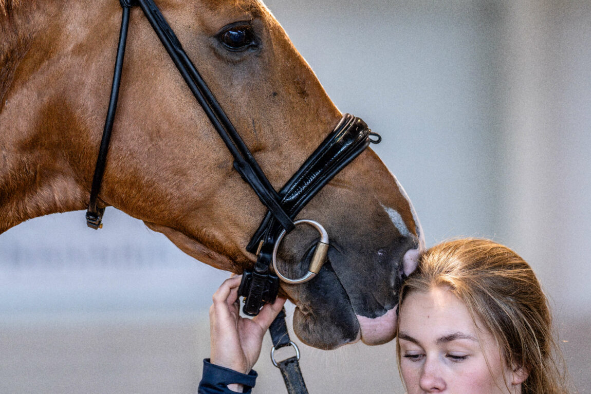 Kristine held fast i grima til hesten mens ho ser ned i bakken. Ho har hovudet sitt heilt inntil mulen på hesten, som ser rett fram over hovudet på ho. 