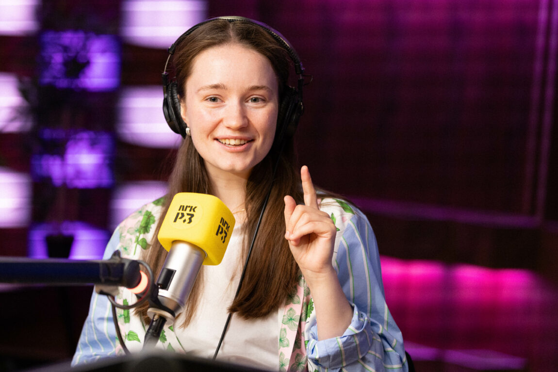 Sigrid holder en finger opp mot taket. Hun sitter i radiostudio og småsmiler. 