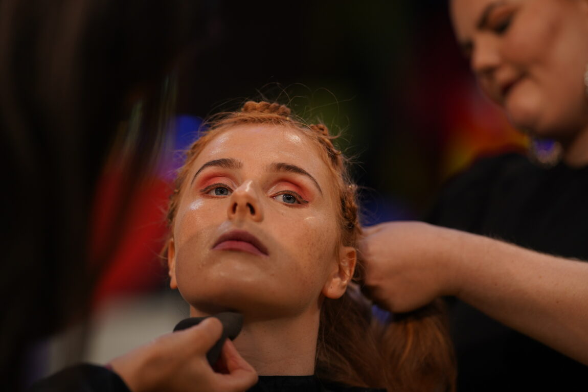 Anna blir sminket. Hun har et strengt blikk, rosa øyenskygge og svart eyeliner. I bakgrunnen ser du at en sminkør og frisør fikser håret og ansiktet.