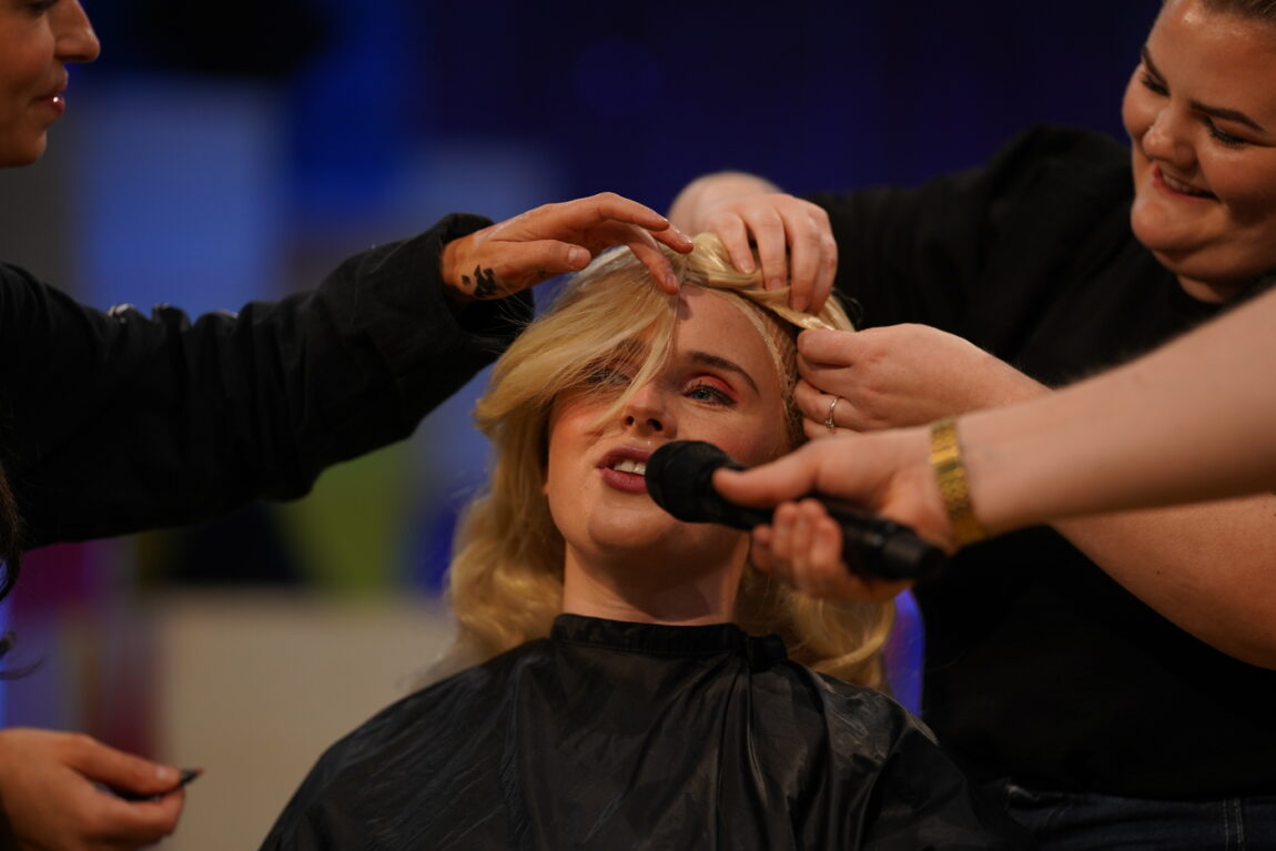 Anna blir sminket i sminkestolen i TV-studio. Sminkør og frisør fikser på den blonde parykken som festes på håret. Anne er ferdig sminket med rosa øyenskygge, svart eyeliner og rosa lepper.