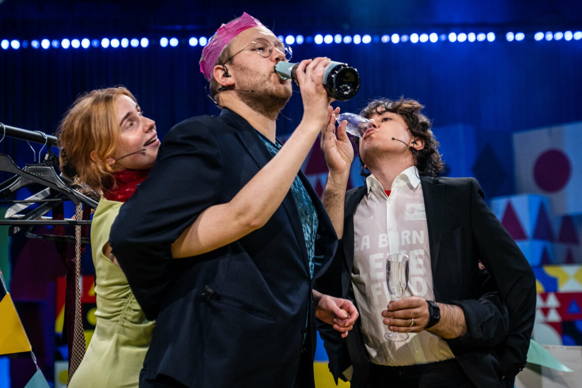 Anna står bak Egil, og holder en flaske opp mot munnen hans. Anna er ikledd en grønn kjole og et rødt sjal, mens Egil er ikledd en rosa krone. Henning drikker fra et champagneglass som Galvans hender holder.