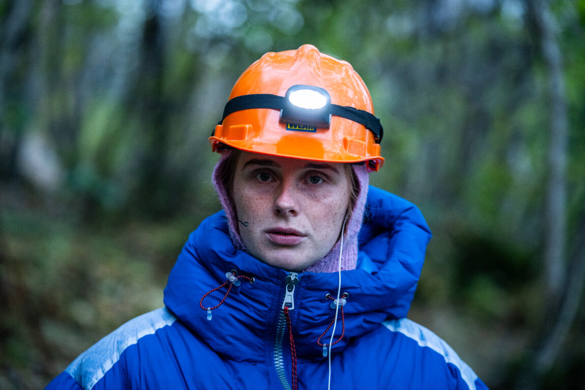 Anna er i en skog. Hun har på seg blå boblejakke, votter og oransje hjelm med hodelykt.