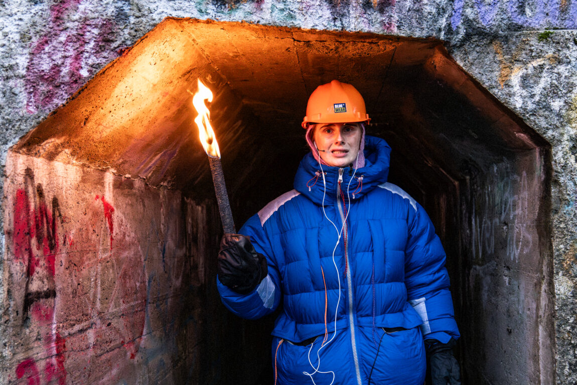 Anna står i åpningen av en grotte. Hun holder en brenennde fakkel. Hun har på seg blå boblejakke, svare votter og en oransje hjelm.