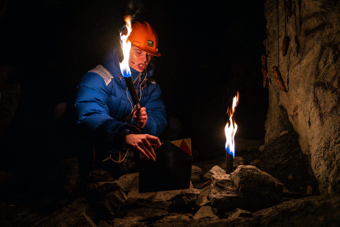 Anna sitter i en mørk grotte. Hun har på seg en blå boblejakke, ornasje hjelm, og holder en brennende fakkel. Hun holder en et brev i den andre hånden.