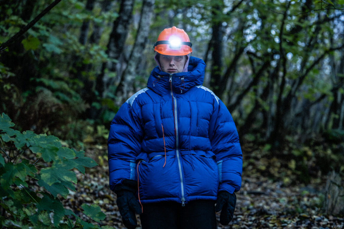 Anna er i en skog. Hun har på seg blå boblejakke, votter og oransje hjelm med hodelykt.