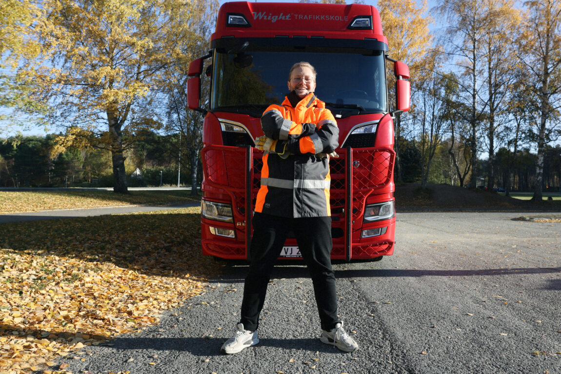 Egil står forran en stor, rød lastebil. Han har på seg en oransje arbeidsjakke. 