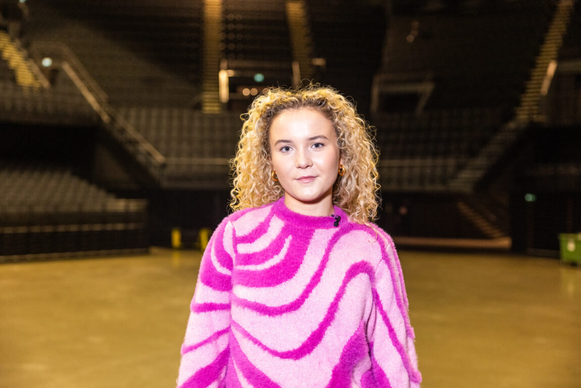Emma Steinbakken ikledd en lyserosa genser med bølgete mønster i neonrosa fuskepels smiler skjevt til kamera. Hun står i Oslo Spektrum foran tomme tribuner.