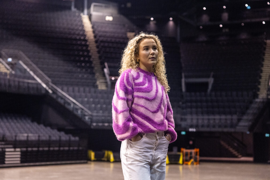 Emma Steinbakken har på seg en blå genser med rosa pels. Hun har hendene i buksa og ser til siden. Bak hun er det masse tomme seter.