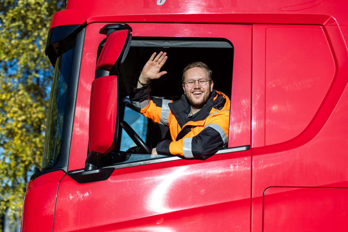 Egil sitter i førersetet i en rød lastebil. Han har på seg en oransje arbeidsdress og ser ut av vinduet fra siden og vinker til kamera.