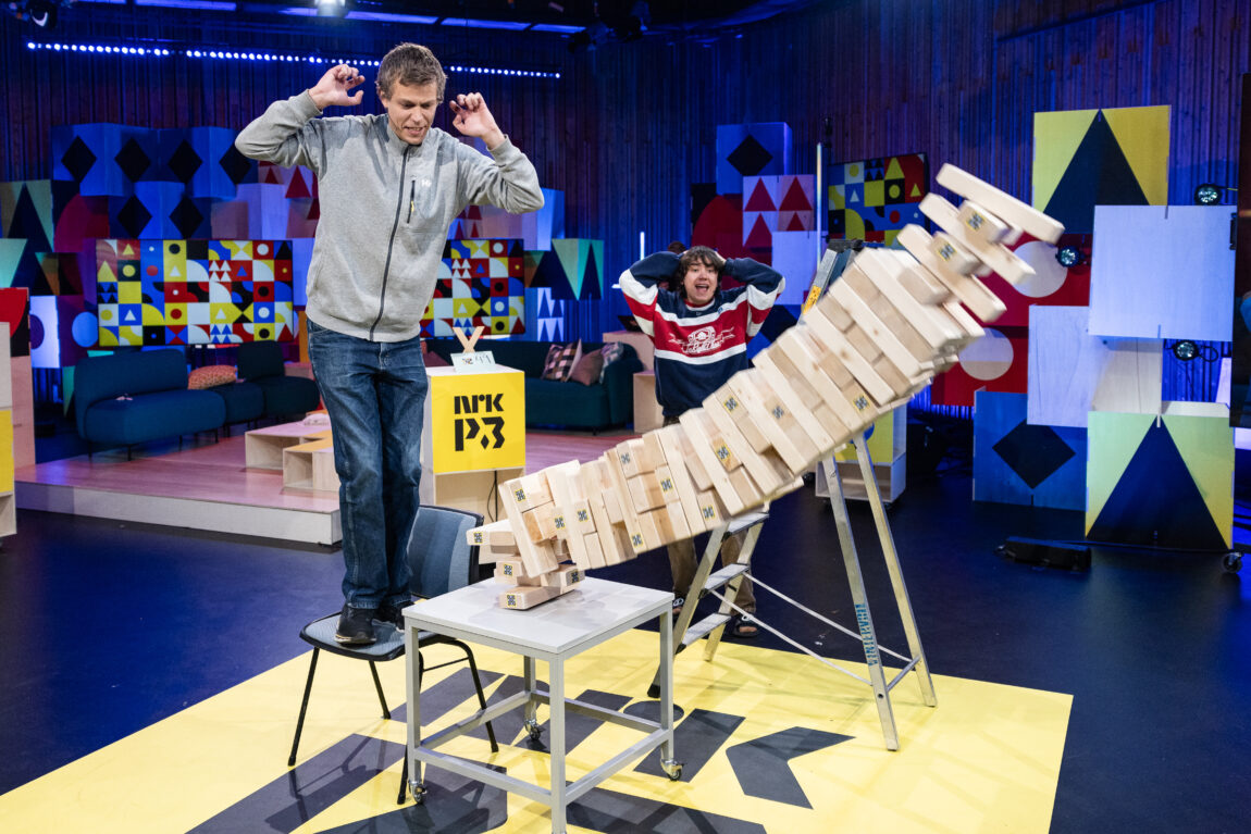 Steinar står på en stol ved siden av et bord med jenga-spill på. Jenga-spillet er på full veg ned mot bakken. Henning står bak og holder seg på hodet.