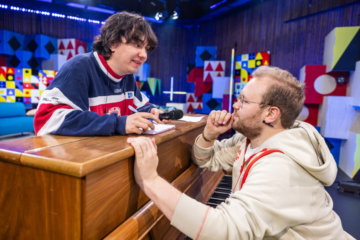 Hennig lener seg over pianoet mens han holder en mikrofon i den ene hånden, og en penn i den andre. Egil sitter på pianokrakken.