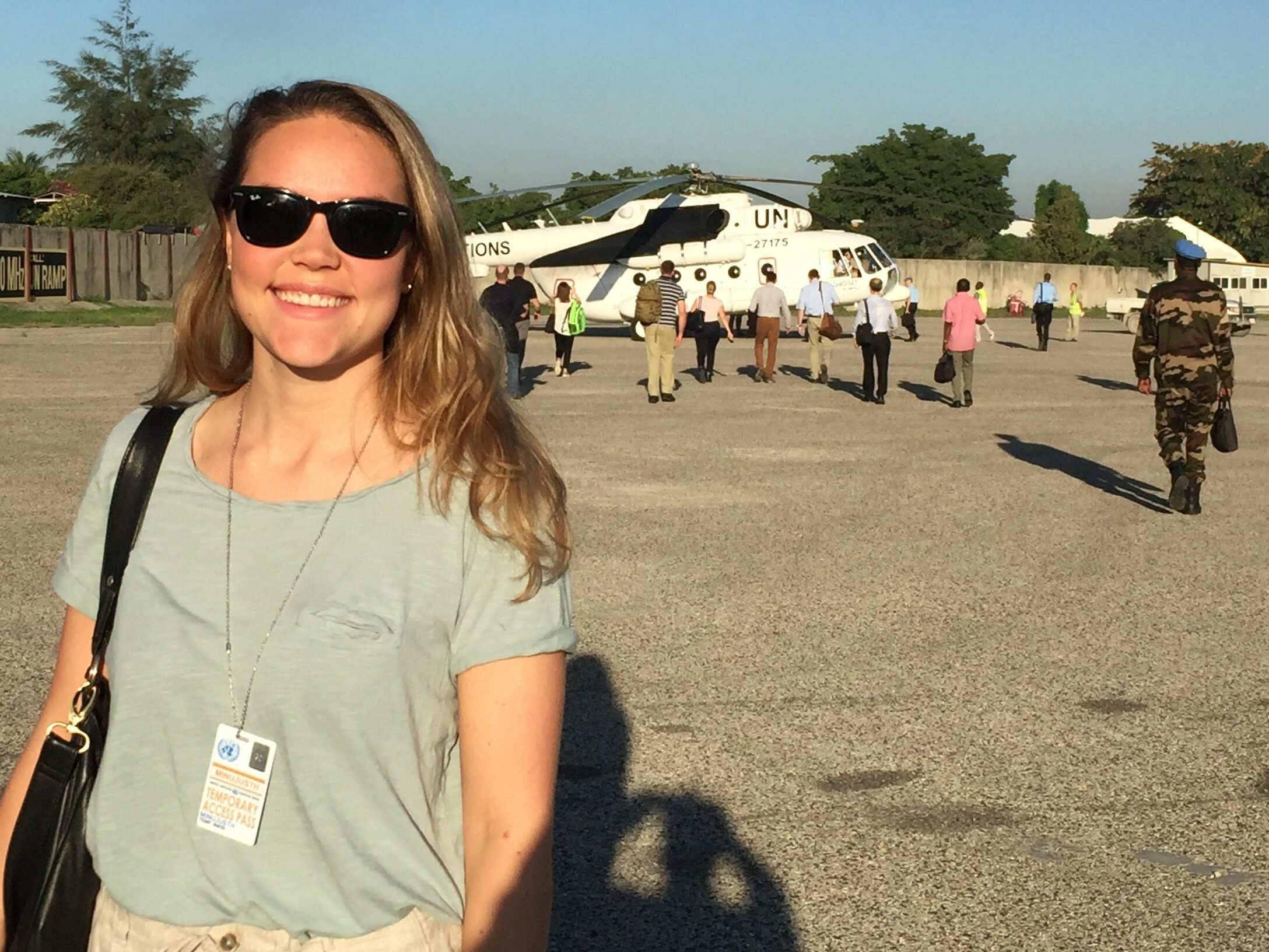 Juni står i solskinnet på ein flyplass, 50 meter framfor eit stort, kvitt helikopter. Bak henne går 14 personar mot helikopteret.