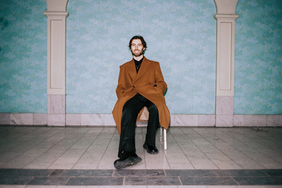 Aden Foyer sitter på en stol med en brun frakk og en svart bukse. Han smiler, har tredagers skjegg og svart midtskill. 