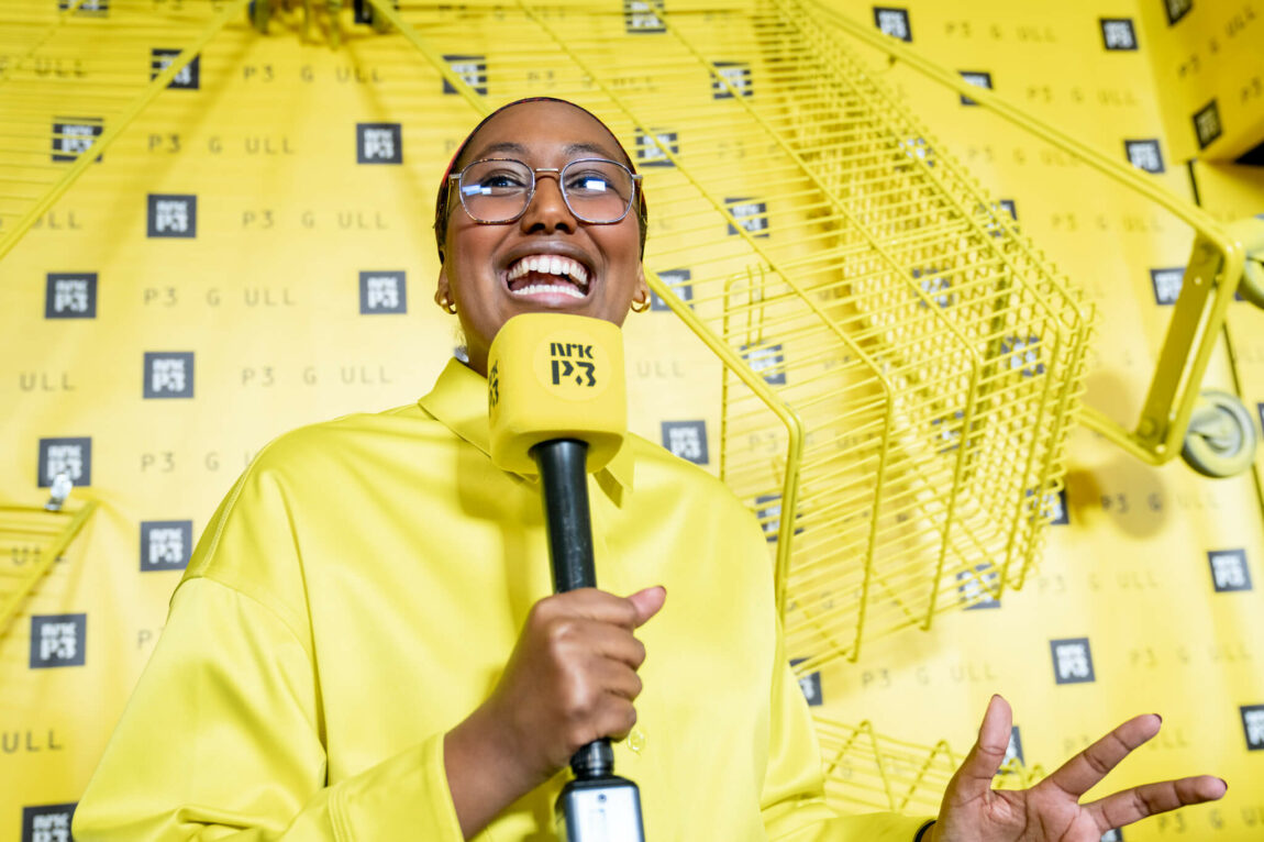 Hani står forran en gul vegg med P3-logo på, og gule handlevogner som er hengt opp. Hun holder en gul P3-mikrofon og er kledd i en gul skjorte. 