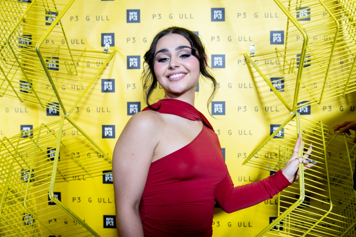 Alessandra står forran en gul vegg med P3-logo på, og gule handlevogner som er hengt opp. Hun har på seg en rød kjole som er langermet på den ene siden og uten arm på den andre. 