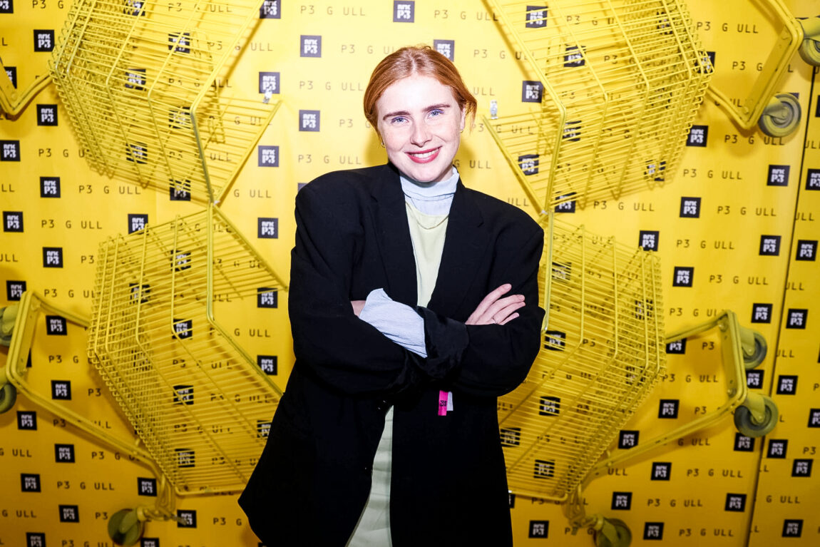 Anna Nor Sørensen står forran en gul vegg med P3-logo på, og gule handlevogner som er hengt opp. Hun har oransje hår satt bak, og hun har svart jakke med en gul kjole under. Armene hennes er i kryss, og hun smiler til kamera. 