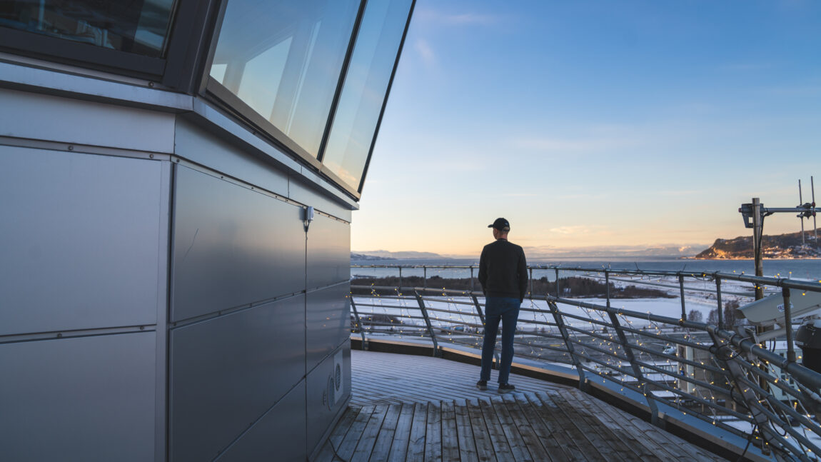 Håkon står med ryggen til kamera på terrassen til kontrolltårnet. I forgrunnen vises flyplassen, snødekket, og en vakker utsikt over Trondheimsfjorden badet i sollys. Det ser veldig kaldt ut.