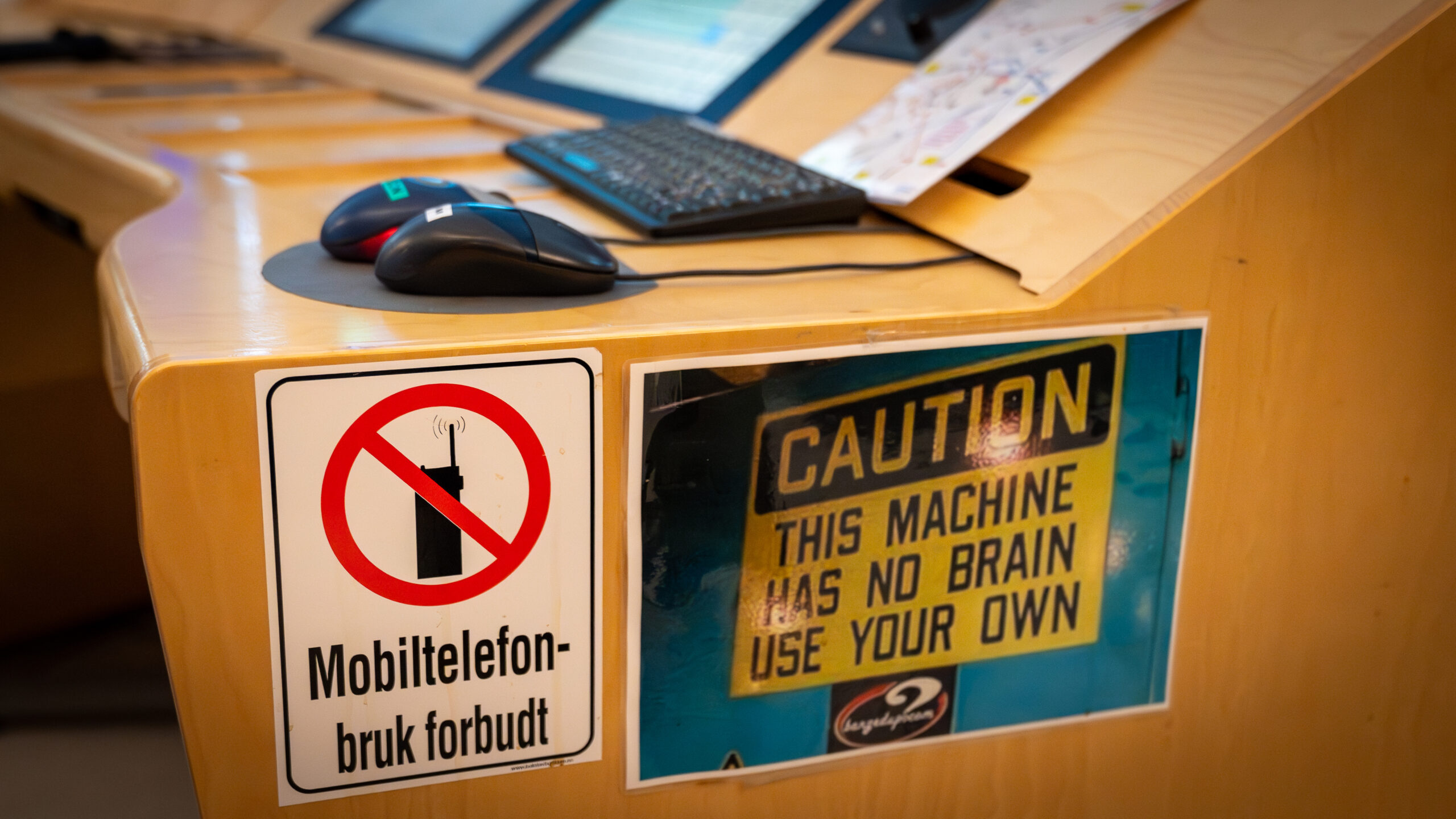 To skilt henger på en pult, på det ene står det: "mobiltelefonbruk forbudt", på det andre står det: "caution, this machine has no brain, use your own"