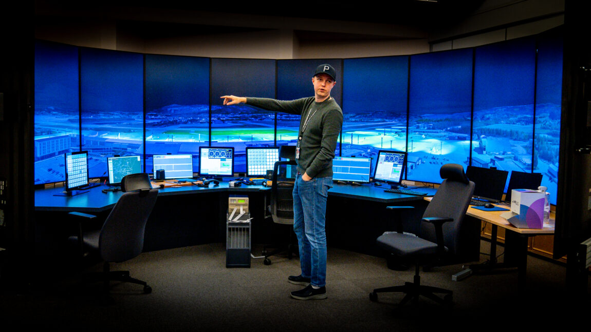 Håkon står foran en simulator med ni store skjermer som viser en flyplass. Foran skjermene er en kontorpult i vinkel, med ni ordinære dataskjermer på. Håkon peker til venstre, og er midt i en forklaring av noe.