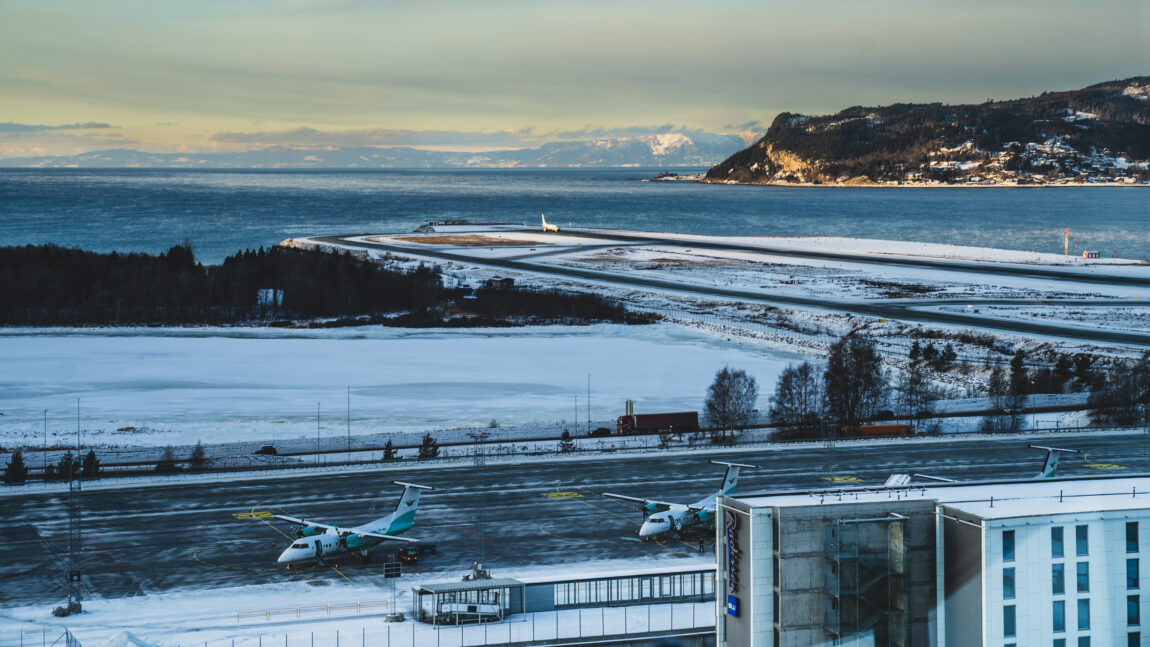 Værnes flyplass i Trondheim, dekket av snø. På rullebanen står flere Widerøe fly parkert. I bakgrunnen er Trondheimsfjorden. Det er nydelig vær.