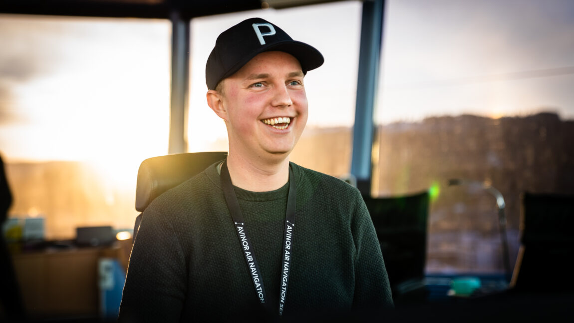 Flygeleder Håkon Kustås er avbildet på jobb i tårnet på flyplassen. Han smiler bredt med åpen munn og tenner, og kikker til høyre for fotografen. Bakgrunnen er en vakker solnedgang. Han har caps, blondt hår, blågrønne øyne og en grønn genser.