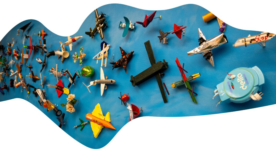 Masse små lekefly, hjemmelagde, er festet på en blå vegg. De er laget av ymse materialer, for eksempel et vaffeljern, en dorull, et påskeegg, lego - flyene er laget av alt mulig.