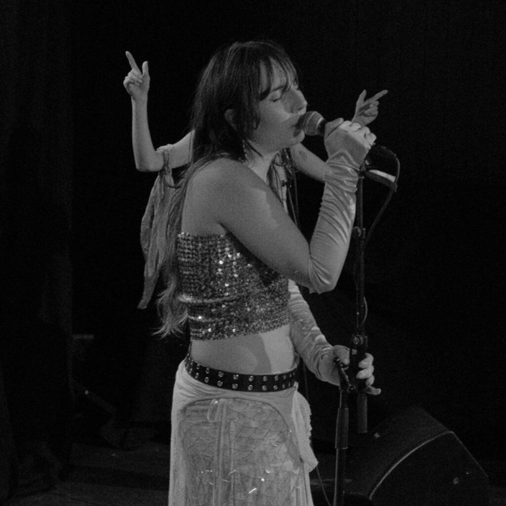 Jez_Ebel synger på scenen i dette svart/hvitt bildet. Hun har på seg noen fancy hansker og holder rundt mikrofonen med øynene igjen.